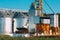 Modern Granary, Grain-drying Complex, Commercial Grain Or Seed Silos In Sunny Summer Rural Landscape. Corn Dryer Silos