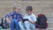 Modern little pupils use gadget sitting on steps of the school with backpacks during recess in open air