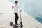 Modern man dressed white shirt and black pants using his phone while standing at a wooden pier with electric scooter