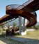 Modern rusted pedestrian and bicycle bridge with flowing Ticino river below. Detail of the spiral staircase and mountain panorama