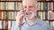 Modern senior man at home talking on cellphone in library. Bookcase bookshelves in background