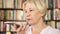 Modern senior woman at home talking on cellphone in library. Bookcase bookshelves in background