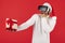 Modern shocked hipster man wears santa claus hat holding gift box and wearing VR headset goggles over red background.