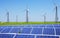 Modern solar station with blue panels and windfarm with wind turbines stand in field with green grass and blue sky above