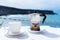 Moka pot espresso maker and cup of coffee on table with coastline and ocean in background