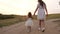 Mom and child hold each other`s hands and walk in the evening in the park. A little daughter in a white dress is walking