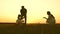 Mom and dad teach a young child to ride a bike at sunset in the park. Teamwork. Silhouette family walk