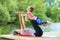 Mom and daughter perform a yoga exercise on the river bank on a warm sunny day