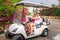 Mom and daughter sit in a golf cart. Family rides an electric car in a beautiful resort. Tourists relax on vacation