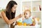 Mom giving homogenized food to her baby son on high chair in kitchen.