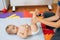 Mom holds the baby in a diaper with a ball in his hands by the legs. Kid lies on a bedspread on top of a colored rug on