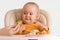 Mom`s hand gives a baby sitting on a feeding chair boiled broccoli