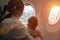 Mom with a small baby in her arms flying on the plane. They look out the window with interest on the earth inside of the airplane