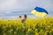 mom with son in her arms, with large satin flag of Ukraine, stand among blooming yellow rapeseed field