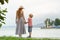 Mom and son standing on the pier and look at each other on the sea background, lighthouse and mountains in the distance.