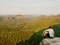 Moment of loneliness. Man in shirt sit on the cliff of rock and watching into forest valley.