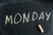 Monday. Name of the day of the week written in yellow chalk on a black chalkboard. Handwritten text. A piece of colored chalk lies