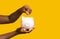 Money Savings. Unrecognizable black woman putting coin into piggybank over yellow background