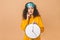 Morning concept. Beautiful shocked amazed curly young woman posing over beige background with clock and sleeping mask