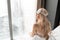 Morning in hotel room. Young woman sits on comfortable bed in mask for sleeping on head. Window of a skyscraper on