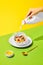 Morning vibe. Female hand pouring milk into bowl with sweet muesli, cereal with berries against green yellow background