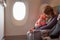 Mother carry her infant baby during flight. Sitting together near the window in airplane