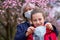 Mother and daughter with a face mask are in the city outdoor, blooming trees, spring season, flowering time - concept of allergies