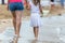 Mother and daughter girl walking together on sand beach in sea water in summer with bare feet in warm ocean waves