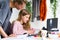 Mother and daughter during homeschooling. Young, blond girl in pink sweater is smiling and asking her mother to help her while