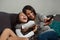 Mother and daughter laugh while watching TV in each other`s arms. The young mother controls the remote control while watching a