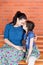 Mother and daughter  in similar clothes hug and sit on the bench in front of red brick wall