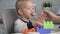 Mother Feeds Porridge With Spoon To BabySitting In Child Chair And Play With Toy