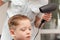 Mother with a hairdryer in her hand in a light blue dress is doing her son`s hair at home during the second period of quarantine.