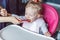 Mother hand feeding infant girl with spoon in high baby chair in kitchen at home