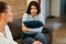 Mother having serious conversation with her teen daughter, sitting together on sofa, lady hugging pillow, looking at mom