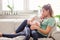 Mother with her daughter sitting together and having good time in the living room in front of window. Two females sitting on the