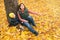 Mother and her little child boy sitting near huge maple tree in autumn park and looking on falling leaves. Fall nature banner with