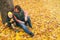 Mother and her little child boy sitting near huge maple tree in autumn park and looking on falling leaves. Fall nature banner with