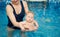 Mother holding her son sitting in her arms in pool and looking in camera. Infant doing water exercises. Health promotion