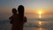 Mother holds baby in her arms while looking at sunset by the sea or ocean shore.