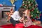 Mother kisses the little daughter on Christmas holiday, sitting near a Christmas tree at home