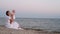Mother in long white dress talking with baby girl sitting on beach. Young mother showing little daughter on the sea.