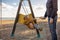 Mother pushing her infant baby boy child on a swing on sandy beach playground outdoors on nice sunny cold winter day in