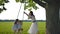 Mother shakes her daughters, on a wooden rope swing tied to an old oak tree.