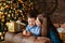 Mother and son hug next to a Christmas tree decorated with golden balls and garlands. The mother kisses the child on the cheek