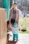 Mother supports her little baby when he do the first steps, smiling woman with toddler child are next to timber house porch