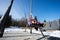 Mother swinging with daughters in big wooden swing in early spring snowy mountains against tiny house and car