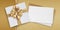 Mothers day gift card, box with golden ribbon bow and white ticket for copy space isolated on beige glittering background, top