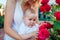 Mothers day. Young woman hugging baby gir in rose garden. Infant looking at flowers touching them discovering world