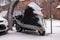 A motorcycle parked on the street, not completely covered with a cover from bad weather, snow lying around, snowfall. Yellow lock